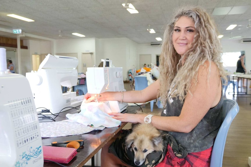 Young woman with long blond hair sits at sewing machine with dog on her lap