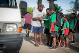 Primary school students are lined up waiting to have their names checked off a list before boarding a community led school bus.