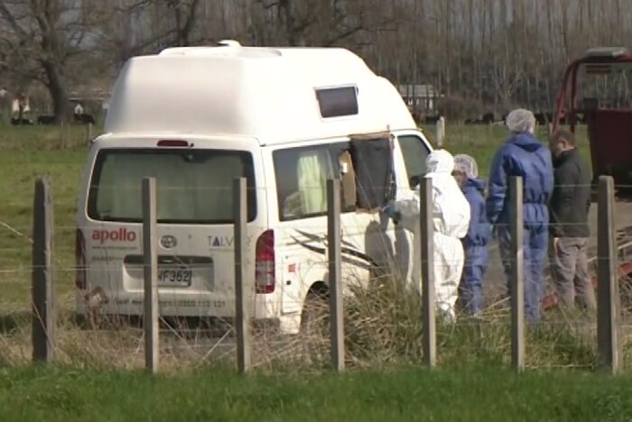 Police dressed in forensic clothing watch on as a campervan is pulled onto a tow truck, on the side of the road.