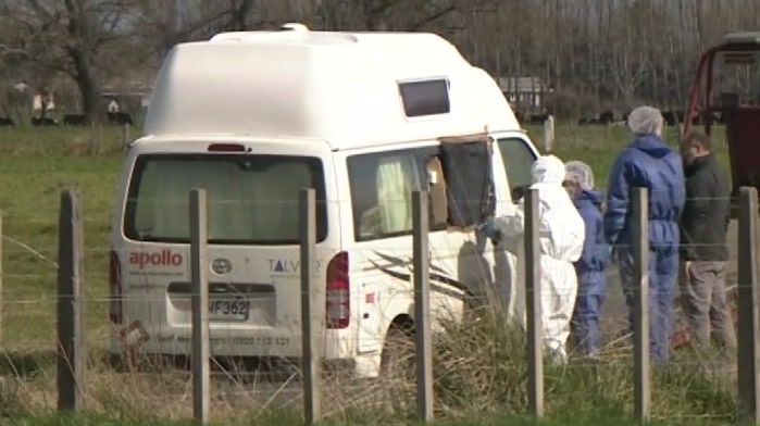 Police dressed in forensic clothing watch on as a campervan is pulled onto a tow truck, on the side of the road.