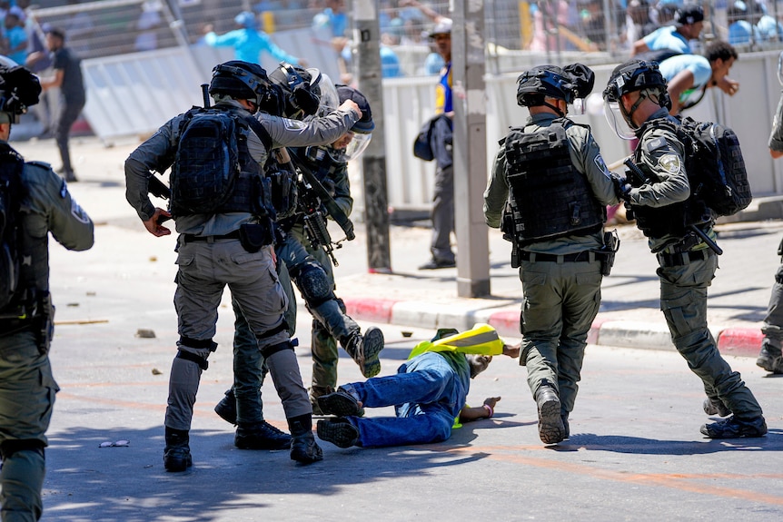 Five police officers in hazard gear surround a man who is on the ground