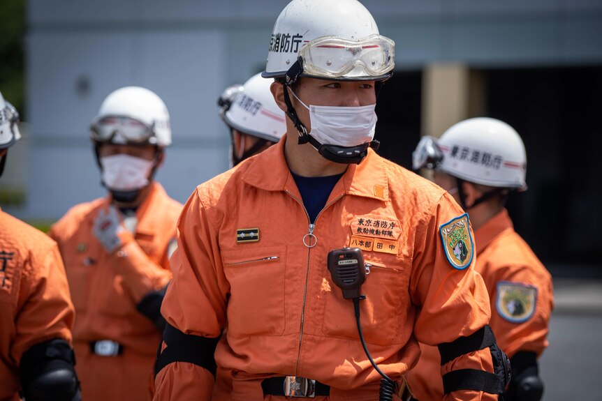 A man in orange jumpsuit and protective gear including helmet and goggles stands among a crowd 