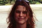 Woman with curly dark hair and black singlet smiles for the camera on a beach.