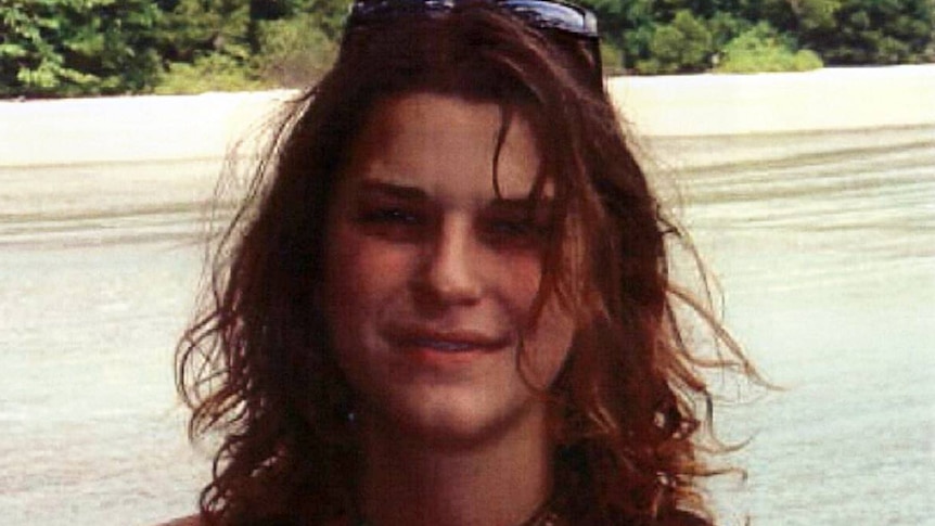 Woman with curly dark hair and black singlet smiles for the camera on a beach.