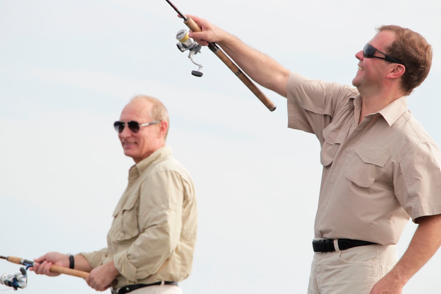 Vladimir Putin and Dmitry Medvedev smiling while holding fishing poles. Both men are in khaki outfits