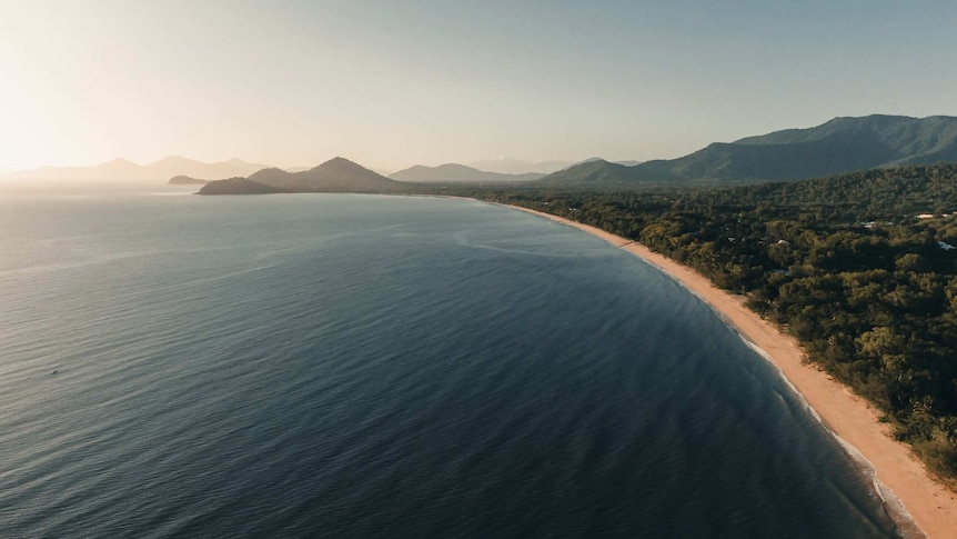 A drone shot of Palm Cove Beach with the sea stretching to meet distant misty hills on the horizon.