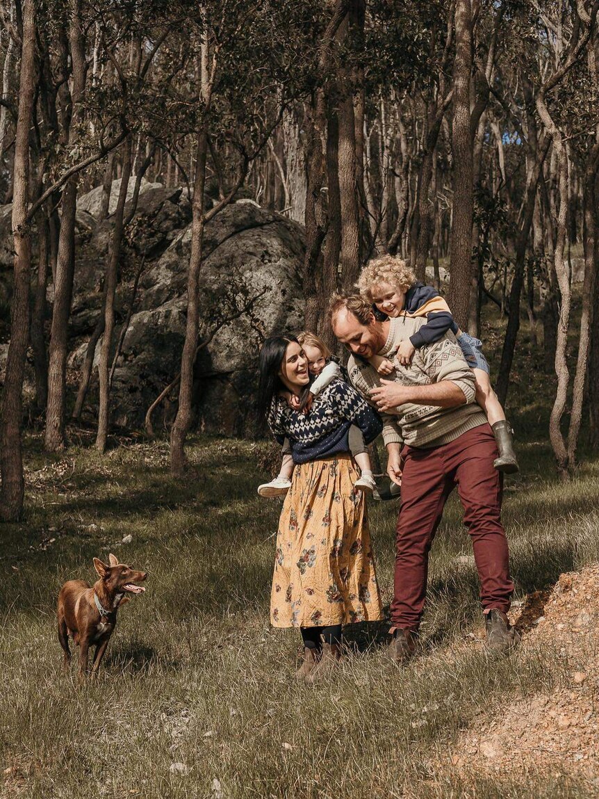 A man and woman each carrying a small child on their shoulders laugh as they walk through the bush with their dog.