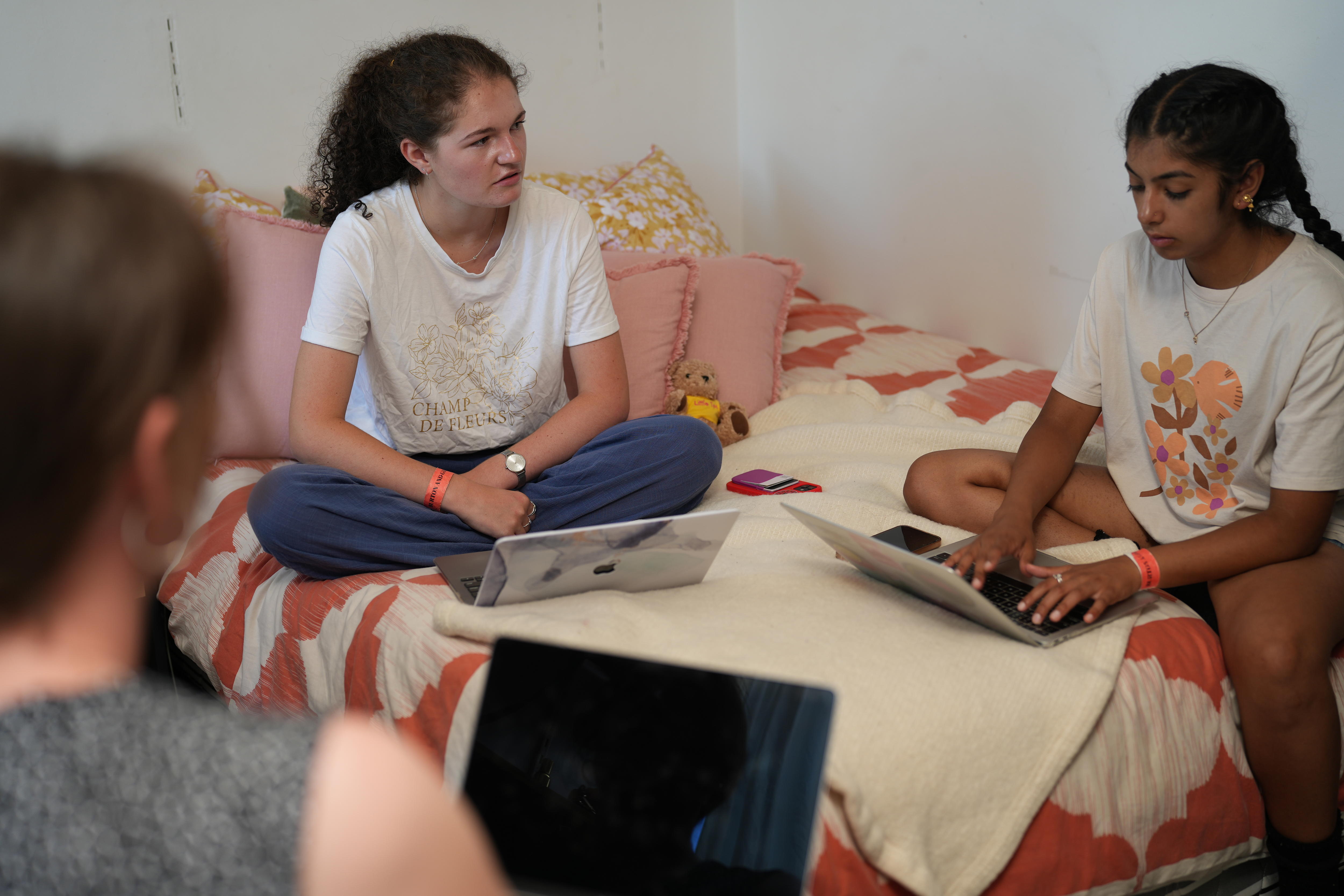 Three teenage girls sit on a bed having a conversation