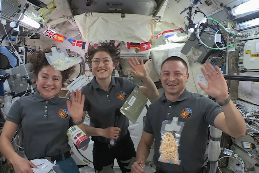 Three smiling, waving people in a space station in zero gravity