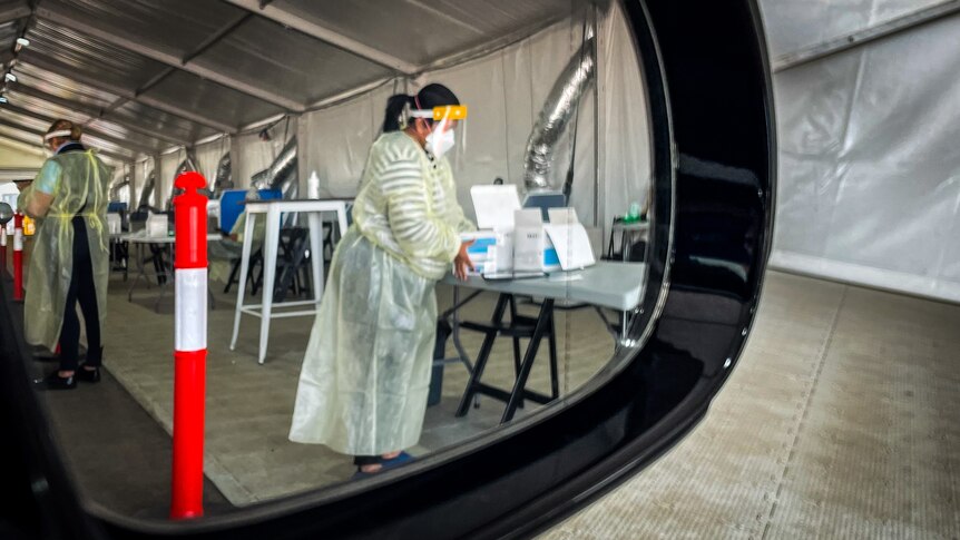 A woman in full PPE works at a table in a tent outdoors, viewed through the side mirror of a car door.
