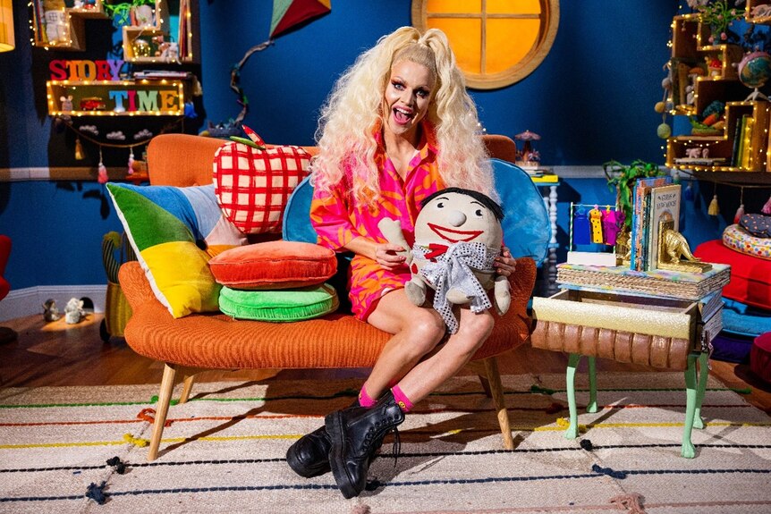 Courtney Act in a long blonde wig and full drag makeup sits on a colourful couch with Play School toy Humpty Dumpty