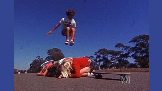 A teenager jumps over a row of kneeling teenagers and towards a skateboard at the end