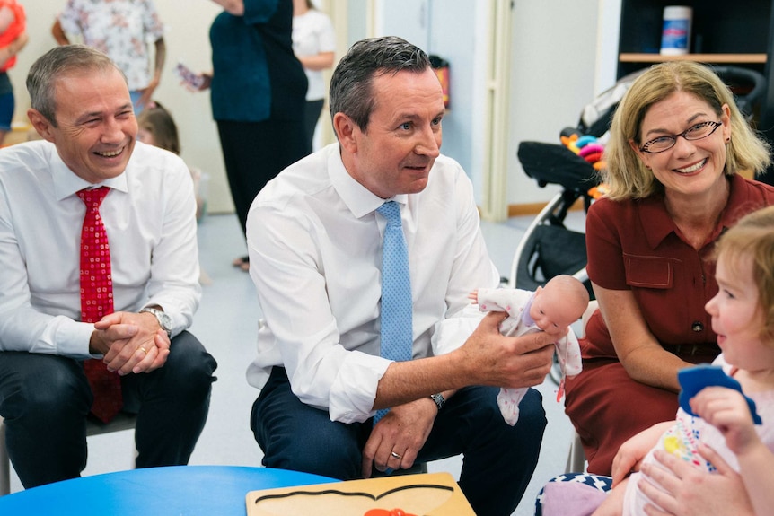 Mark McGowan holds a baby doll and talks to a child.