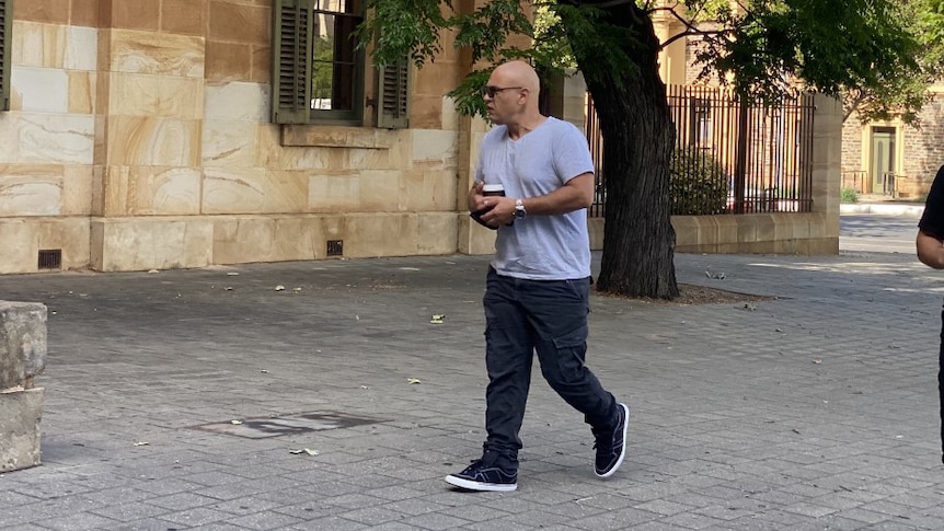 A bald man wearing a grey t-shirt, face mask, sunglasses and black cargo pants walks outside the Adelaide Magistrates Court