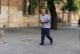 A bald man wearing a grey t-shirt, face mask, sunglasses and black cargo pants walks outside the Adelaide Magistrates Court