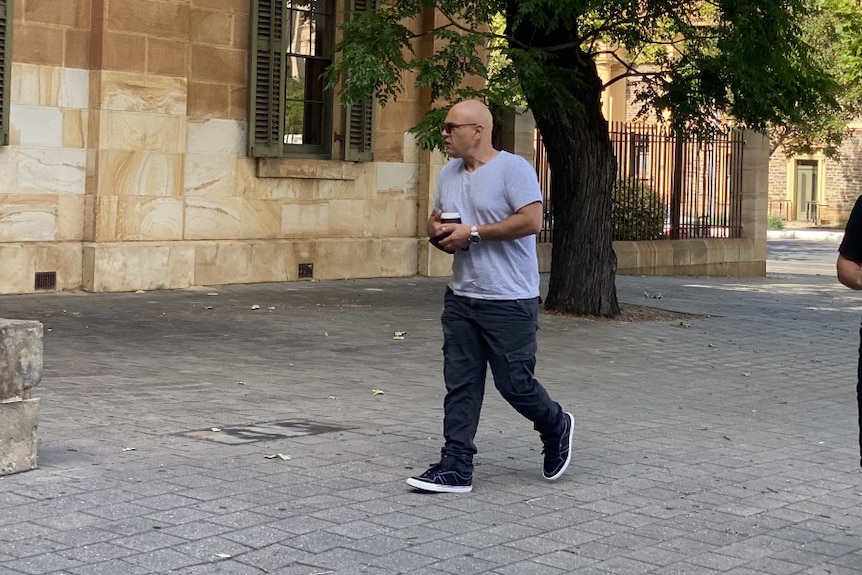 A bald man wearing a grey t-shirt, face mask, sunglasses and black cargo pants walks outside the Adelaide Magistrates Court