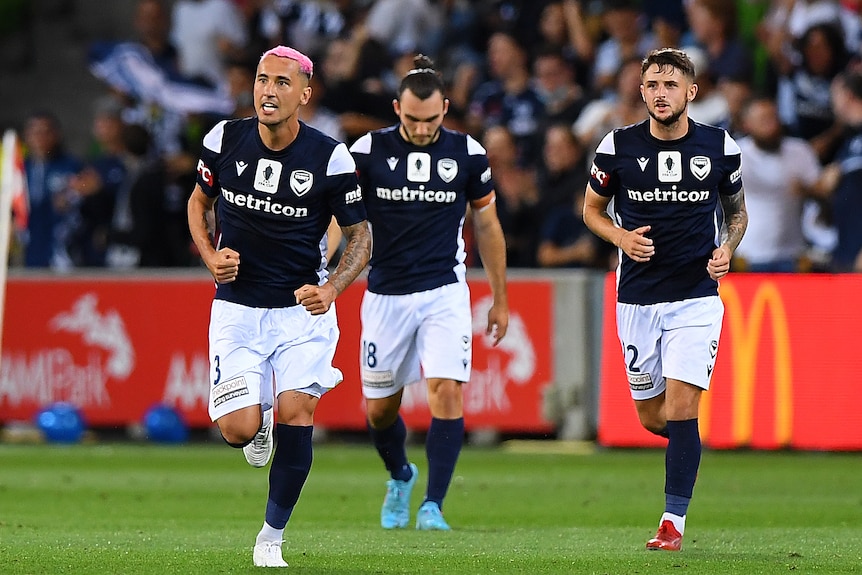 A soccer player in dark blue jersey and pink dyed air looks jubilant as he runs with his teammates on pitch 