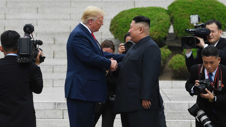 Donald Trump and Kim Jong-un are surrounded by cameramen as they shake hands in front of concrete steps in North Korea.