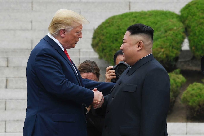 Donald Trump and Kim Jong-un are surrounded by cameramen as they shake hands in front of concrete steps in North Korea.