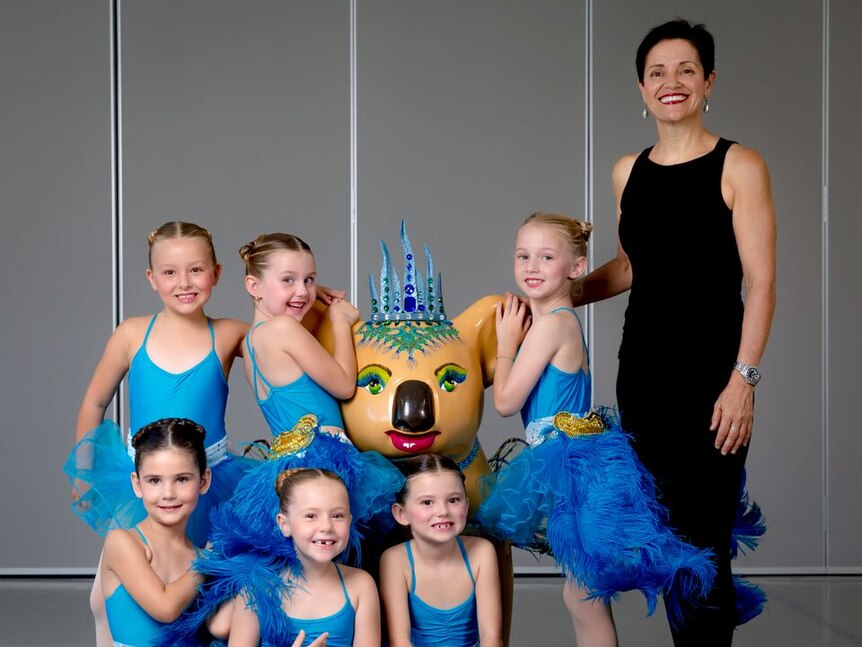 A dance teacher wearing black pants and a black top stands with young dancers in blue ballet costumes, with a koala sculpture.