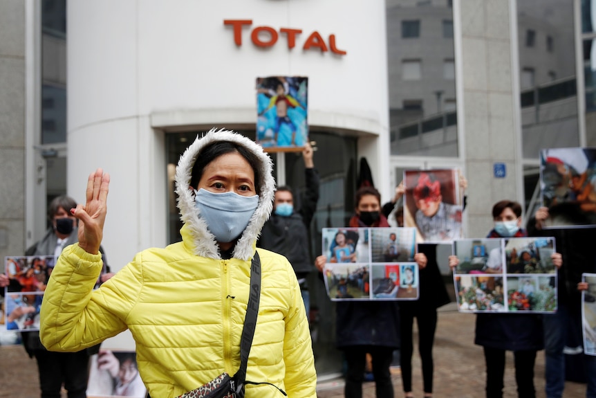 A face-masked Asian woman in yellow jacket holds three-finger saluted in front of protesters at Total building.