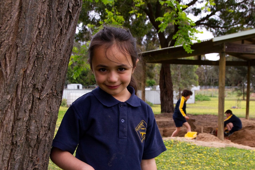 A young girl standing under a tree with two kids playing in the background.