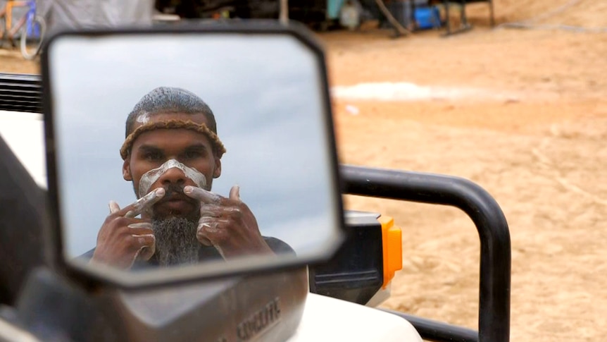 a man puts white traditional face paint on while looking in the side mirror of a wagon