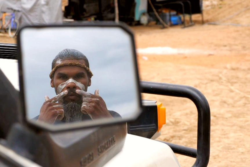 a man puts white traditional face paint on while looking in the side mirror of a wagon