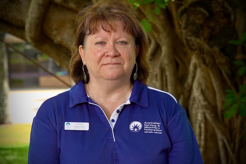 White woman with red hair and blue shirt stands in front of tree