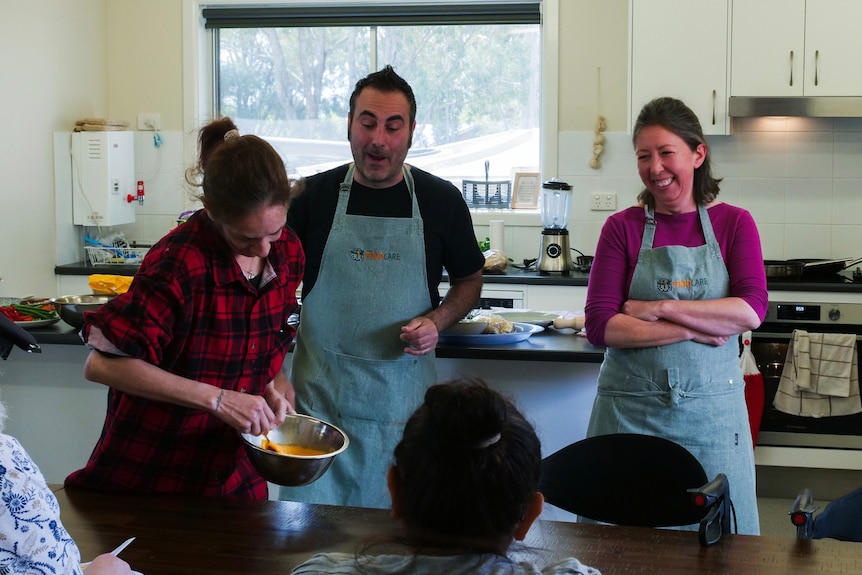 A man and a woman wearing aprons smile watching a woman whisk an egg.