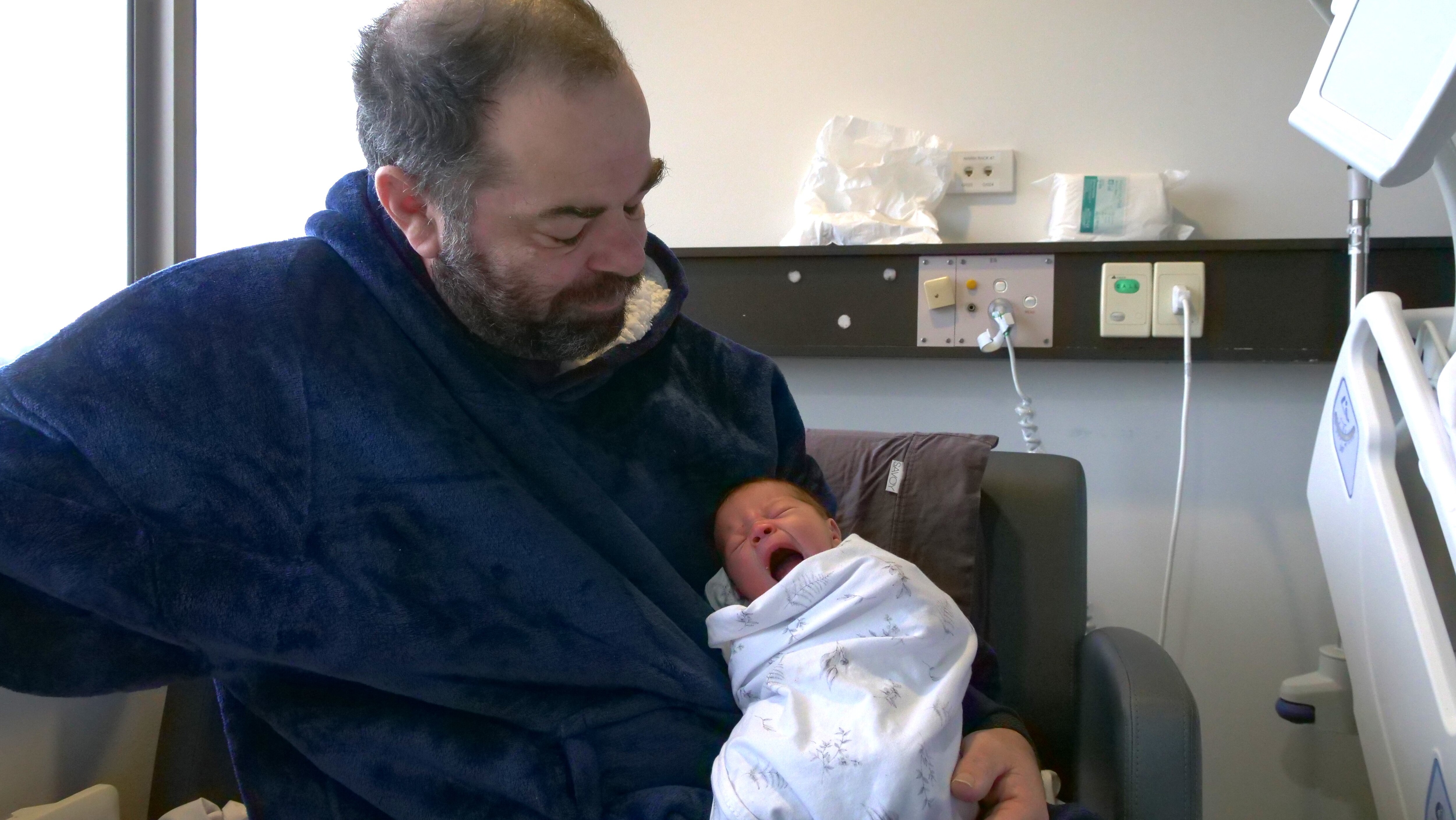 A man in a fluffy navy blue oodie and a baby in a wrap sit on a hospital bed.