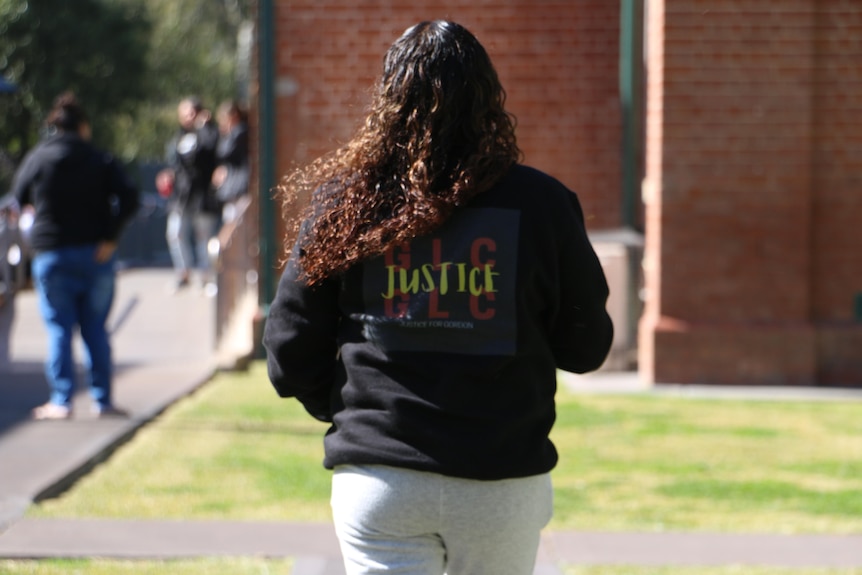 A woman, seen from behind, wearing a black jacket with justice written in yellow and the initials GLC in red.