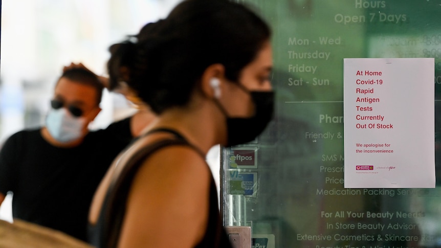 a woman in a mask walking past a sign notifying customers that rapid antigen tests are sold out