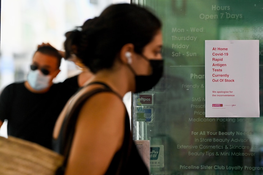 a woman in a mask walking past a sign notifying customers that rapid antigen tests are sold out