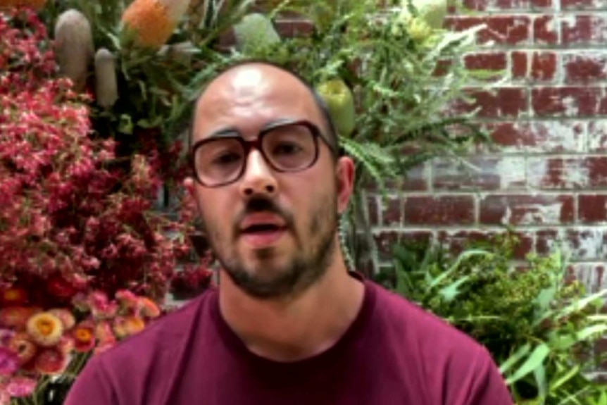 A man in glasses with flowers behind him.