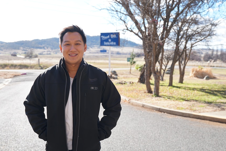 A man in a black jacket stands in a driveway with a motel sign and grass in the background.