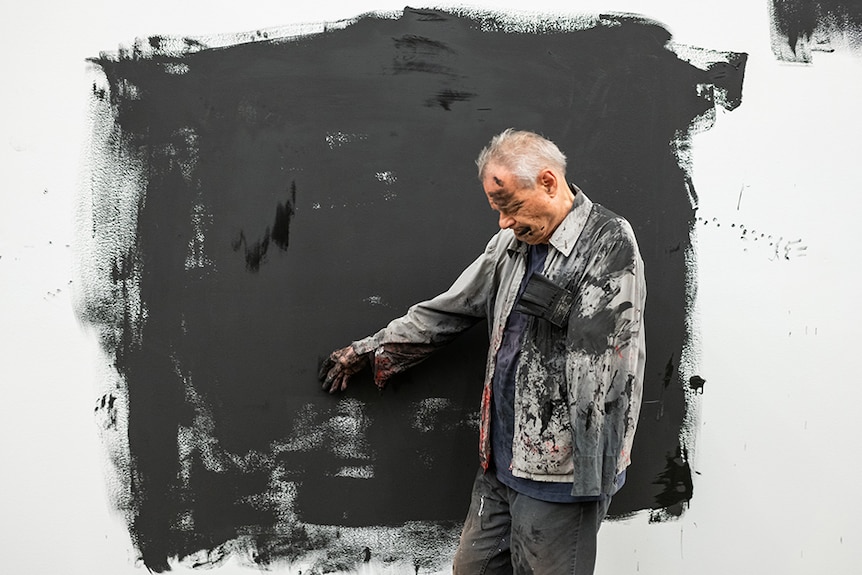 Mike Parr stands in the middle of a roughly painted black square on white gallery wall, his body and clothes covered in paint.