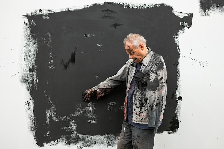 Mike Parr stands in the middle of roughly painted black square on white gallery wall, his body and clothes covered in paint.