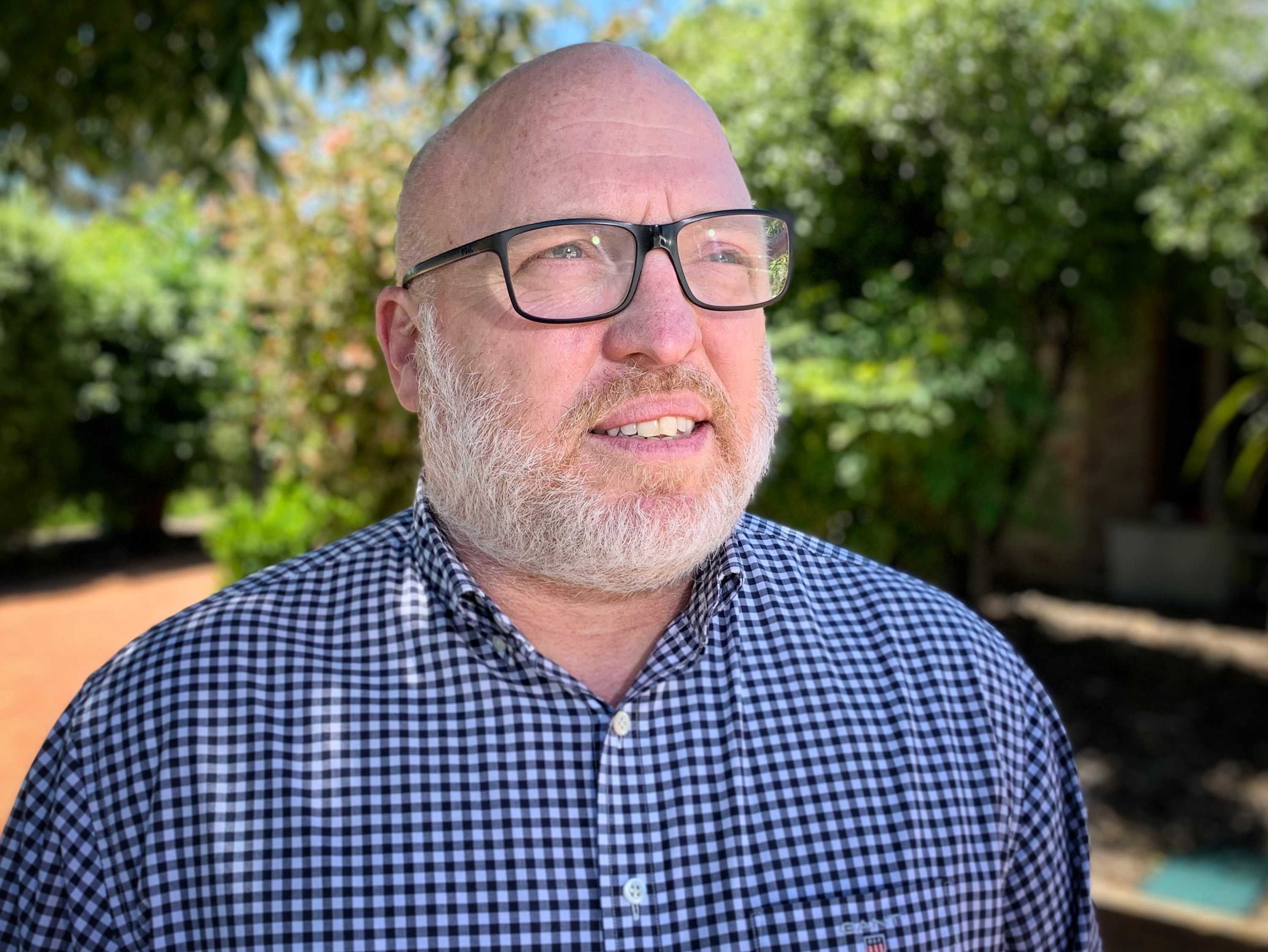A bald man with a beard wearing black glasses and a plaid button-down shirt stares into space.