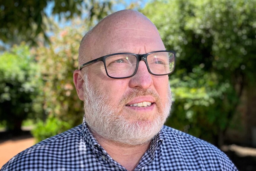 A bald man with a beard wearing black glasses and a plaid button-down shirt stares into space.