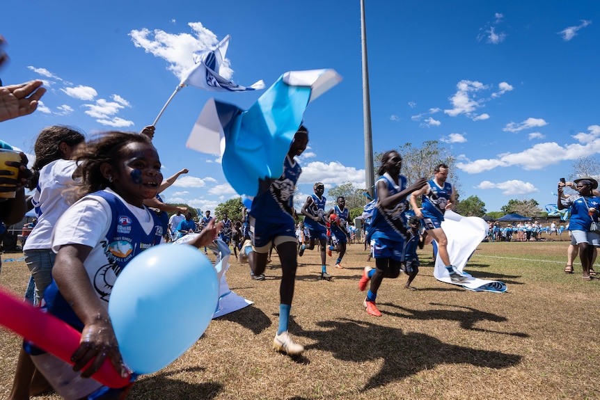 Young men in football jersies run on a football field. Young kids run alongside them holding flags and blue balloons 