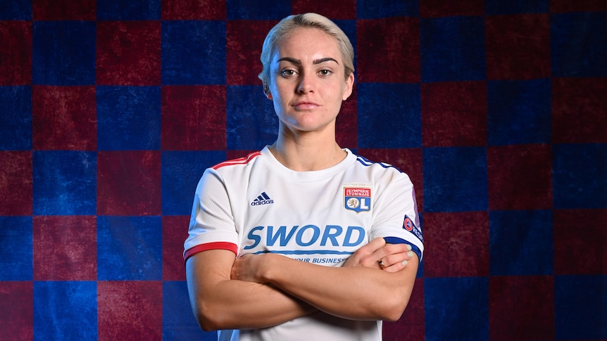 A female soccer player wearing white with a red and blue checked background folds her arms