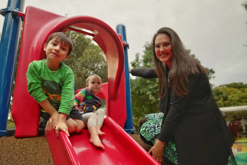A woman smiles as her two small children sit at the top of a slide. 