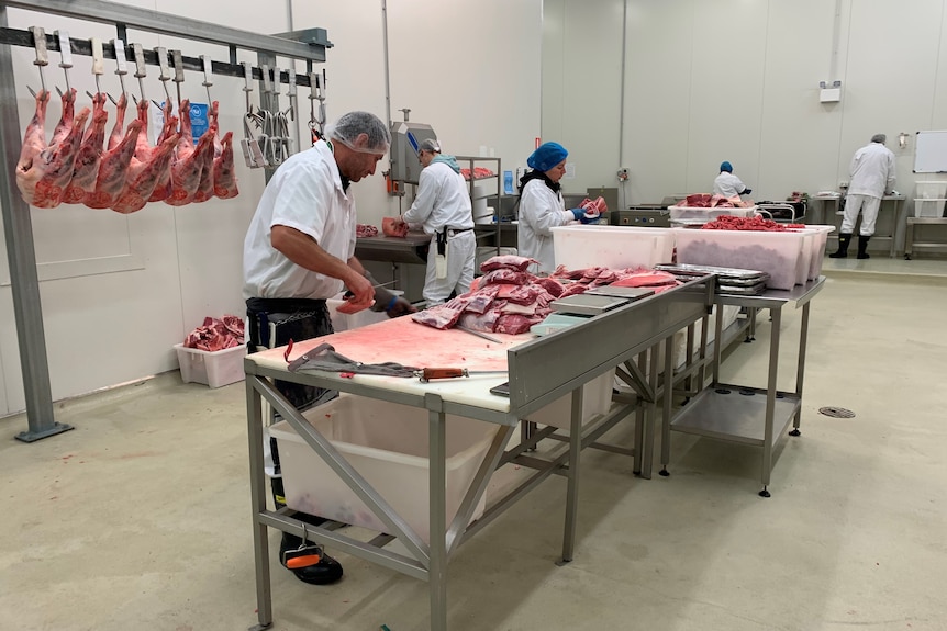 Workers stand around metal tables with cuts of meat piled on them. 