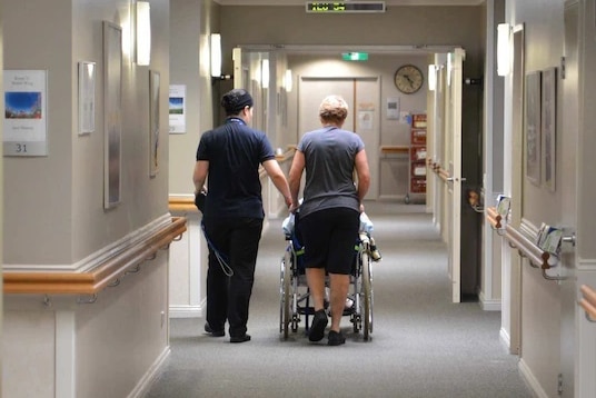 An aged care worker accompanies an elderly resident down a hallway.