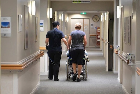 Aged care workers accompany a resident down the hallway of their facility