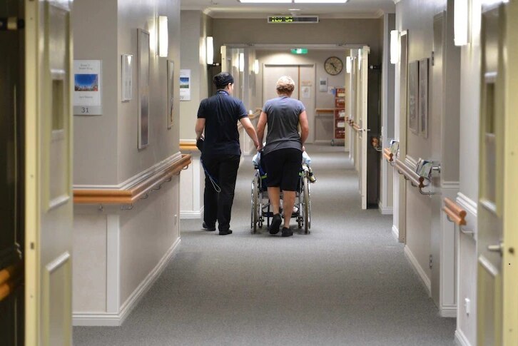 Aged care workers accompany a resident down the hallway of their facility