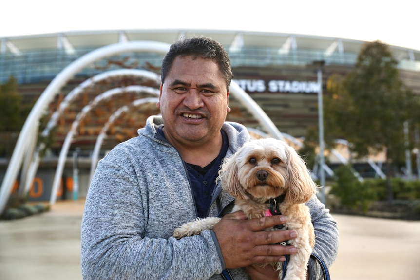 A man is smiling and holding up a small light coloured dog, behind him is a tree and Perth Stadium.
