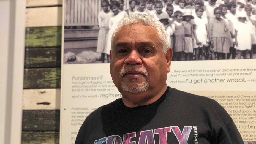 Man with grey curly hair and neatly groomed goatee looks calmly at the camera with a historical photograph in the background