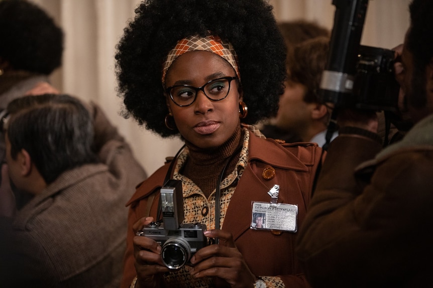 Film still of Kirby Howell-Baptiste as Anita, wearing glasses and holding a camera, surrounded by a press pointing in the other direction.